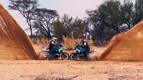 Twee motorrijdsters van het internationale team staan met de voorwielen tegenover elkaar en laten met de achterwielen een indrukwekkende hoeveelheid zand opstuiven.