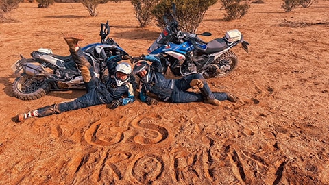 De twee motorrijdsters van het internationale damesteam liggen voor hun motoren in het zand waarin ze GS Trophy hebben geschreven. 